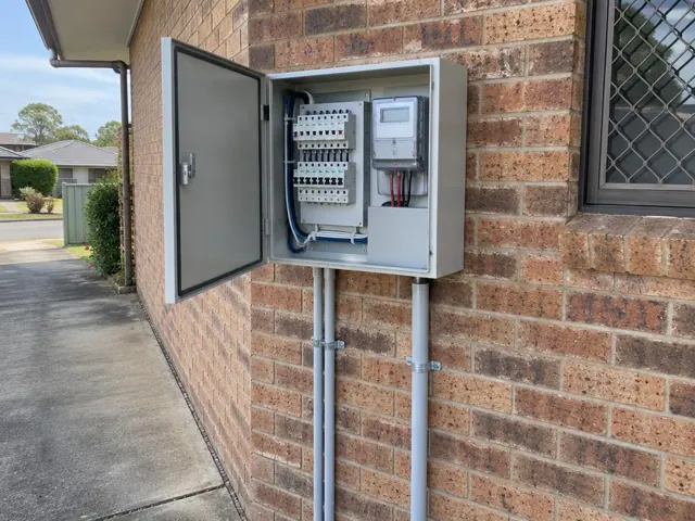 Open outdoor switchboard on a brick wall beside a driveway, with conduits and meter cabinet in Hamlyn Terrace
